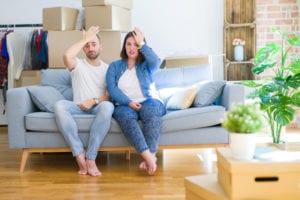 Young couple sitting on the sofa arround cardboard boxes moving to a new house surprised with hand on head for mistake, remember error. Forgot, bad memory concept.